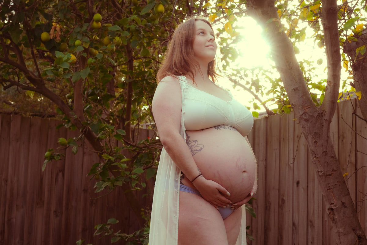Pregnant woman standing outdoors in a garden with trees and wooden fence in the background.